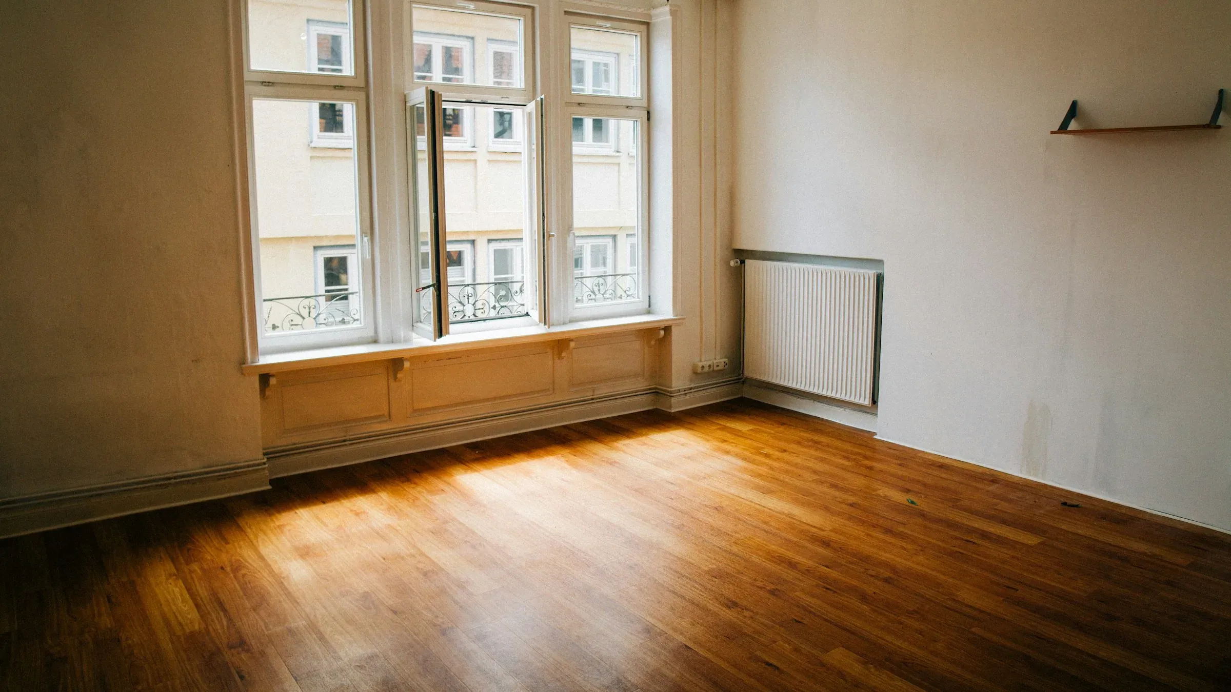 An empty room with a wooden floor and tall windows, cleared except for a small bracket left on the wall.